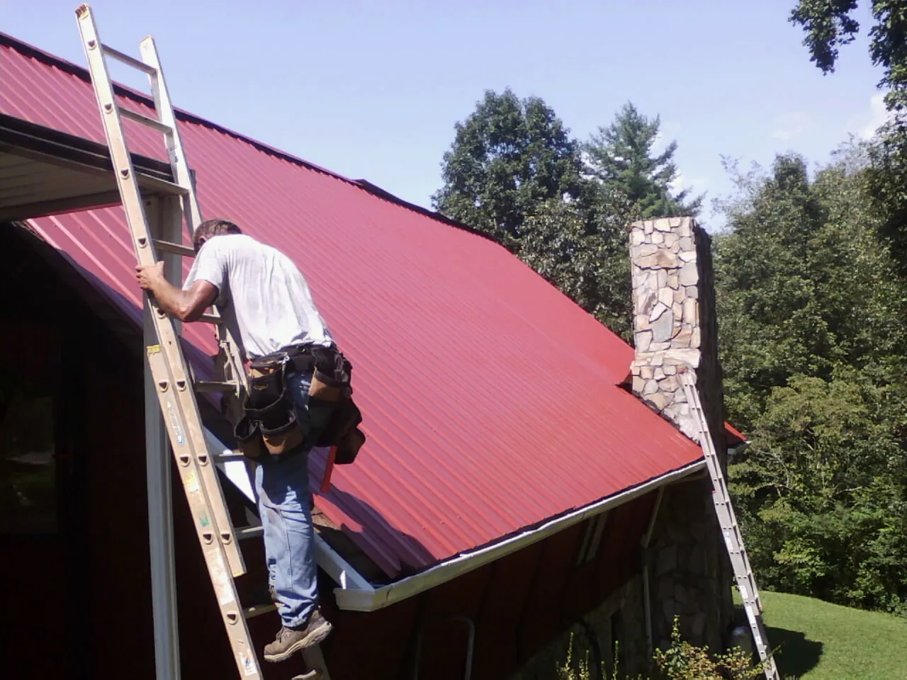 Roofer on a ladder beside a red metal roof and stone chimney — Soffit & Fascia Repair by Roofing Brothers in Minnesota City